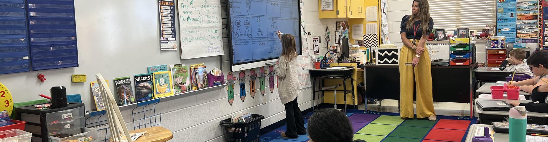 One student works out a problem on the smart board while the teacher and other students look on.