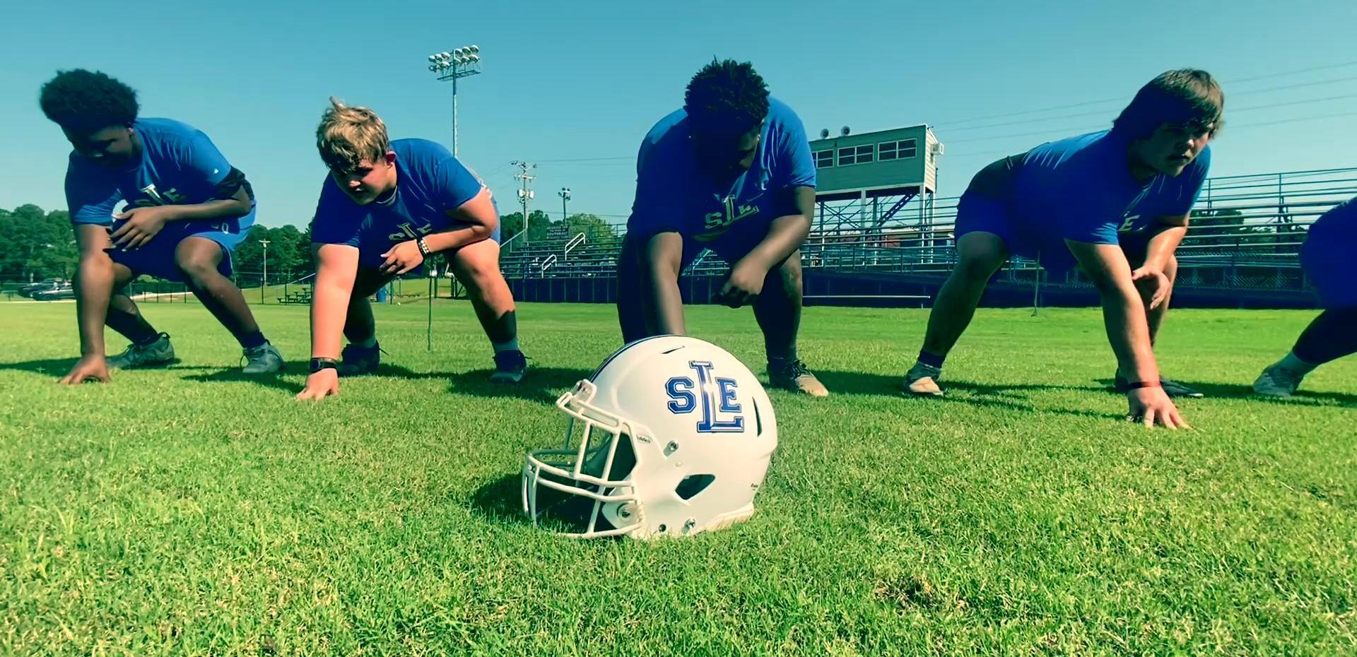 Football players in blue uniforms practicing drills on the field, with a helmet in the foreground.