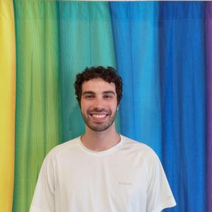 Arco Iris Spanish Immersion School PE teacher Eli Serrao stands in front of colorful yellow, green and blue curtains in a white t-shirt.