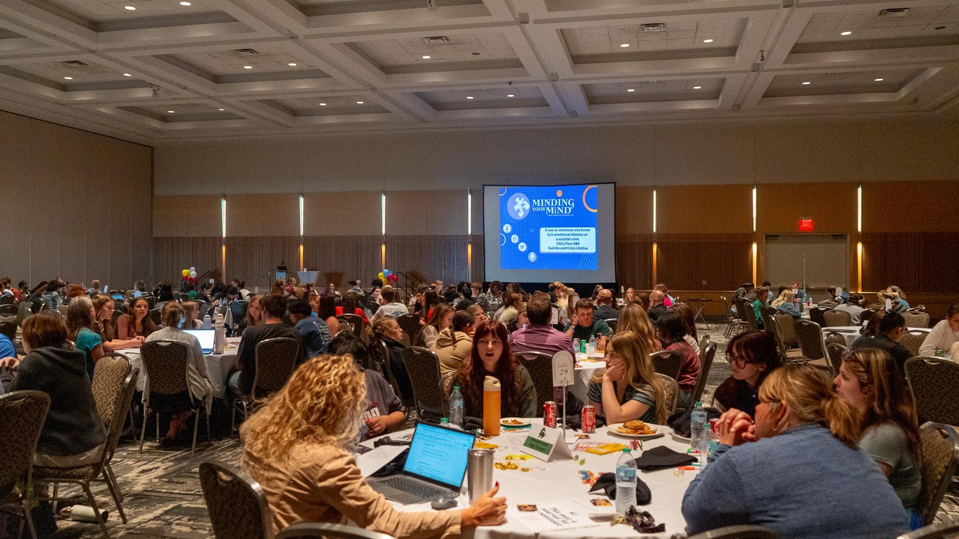 A large conference room filled with students working tables with a presentation screen in the background titled "Minding your mind", during a student mental health symposium.