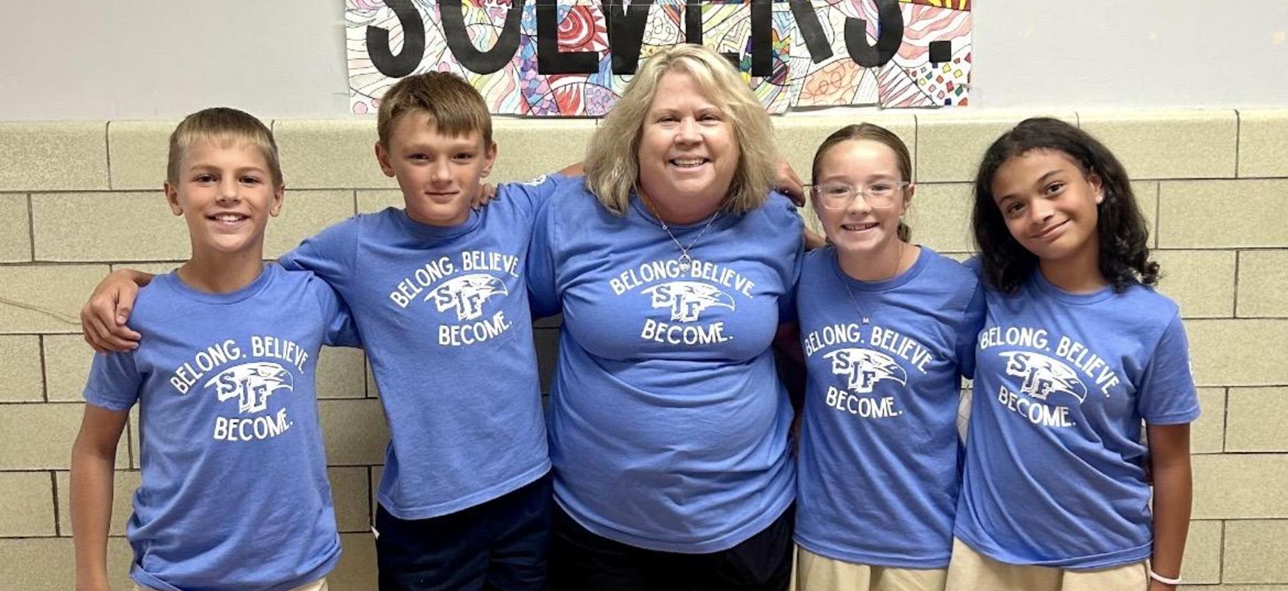 Four children and an adult wearing matching blue shirts, smiling together in a school setting.