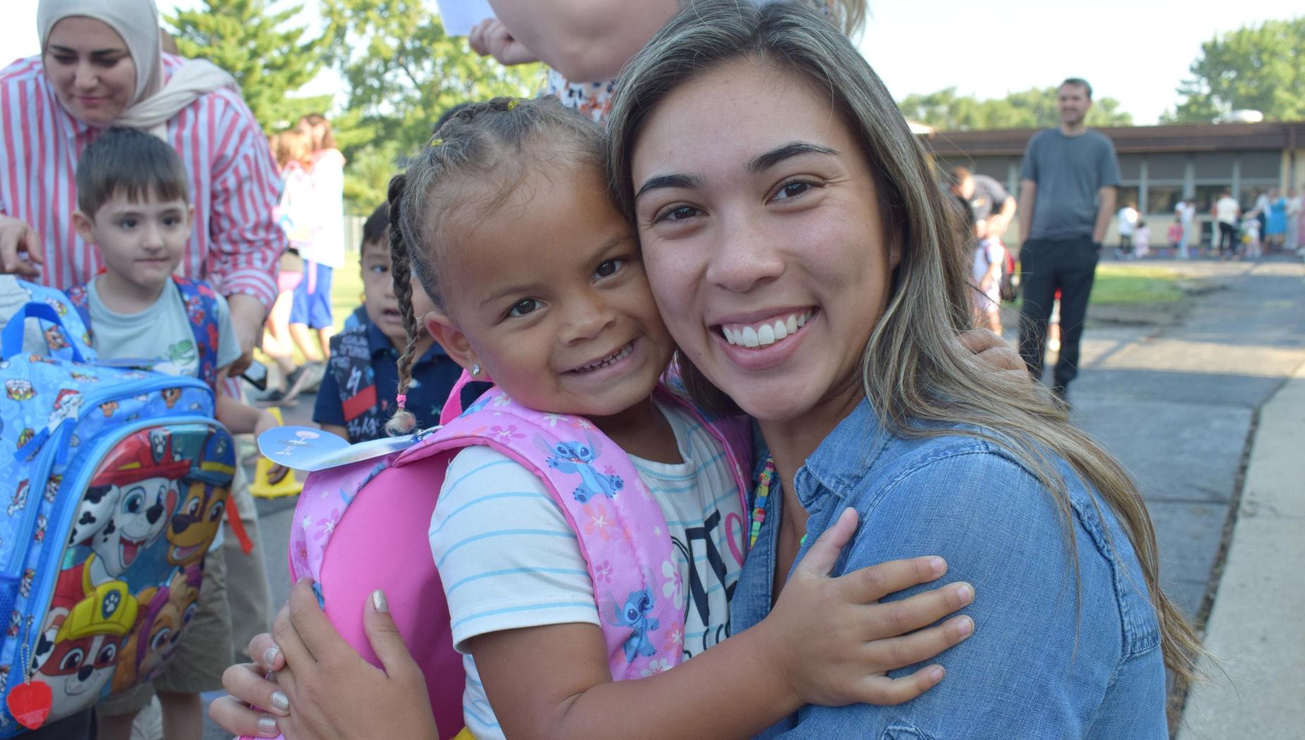 A smiling girl hugging an adult woman outside, both with colorful backpacks.