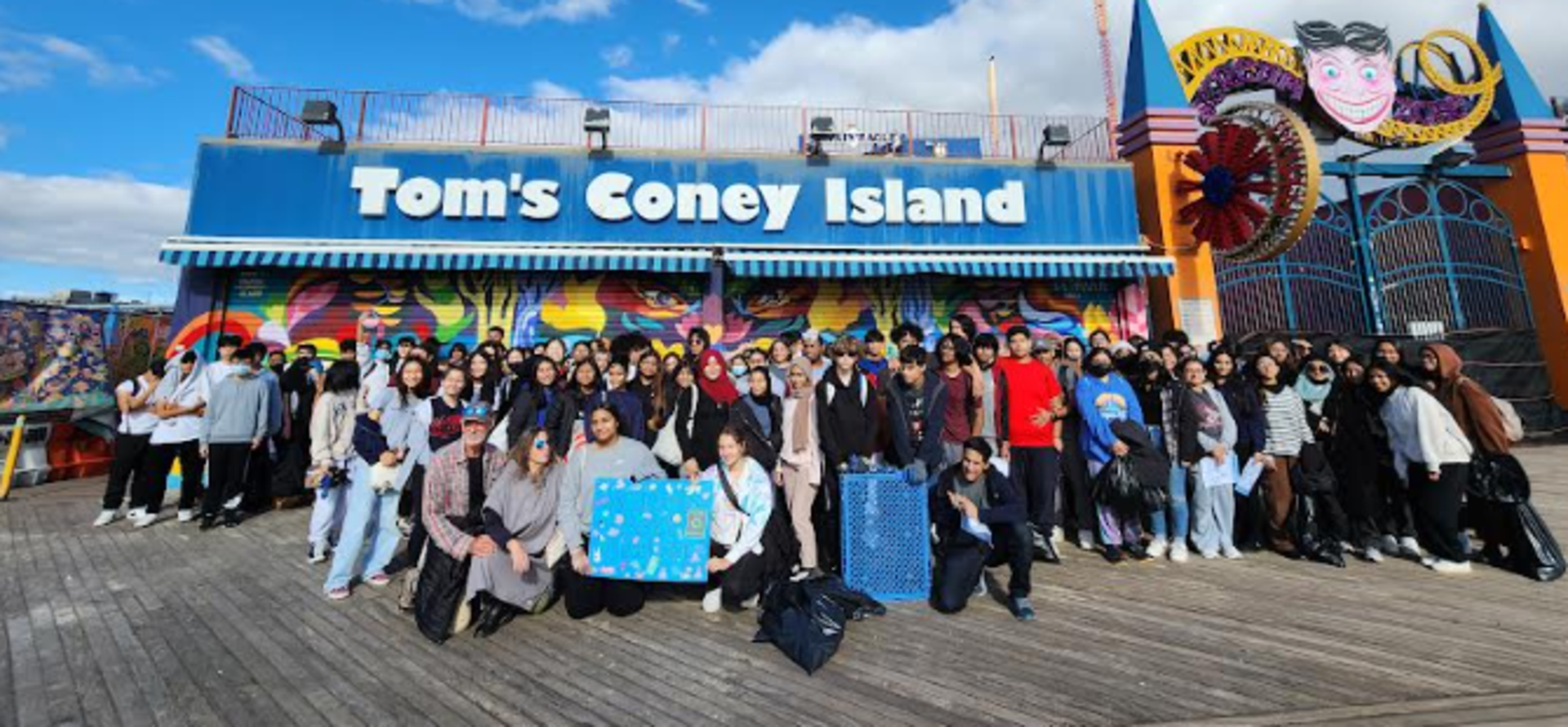 Large group of people posing in front of Tom's Coney Island with colorful background.