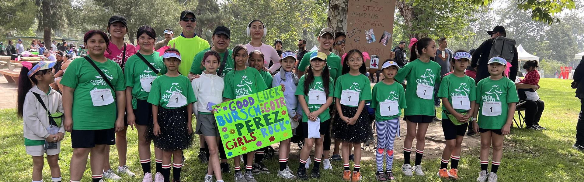 Group of children and adults in green shirts at an outdoor event, holding a sign.