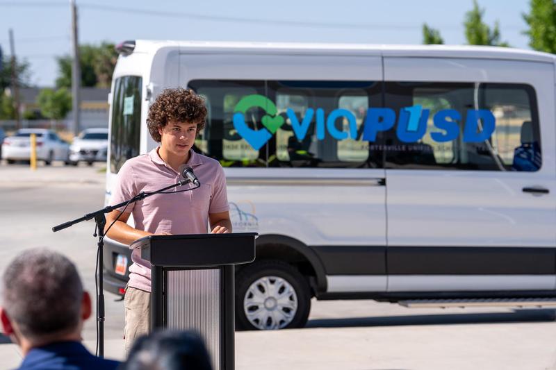 A student speaker stands at a podium addressing the crowd during a ribbon-cutting ceremony for the PUSD and Via Transportation partnership. Behind him, a white electric van displays the Via and PUSD logos.