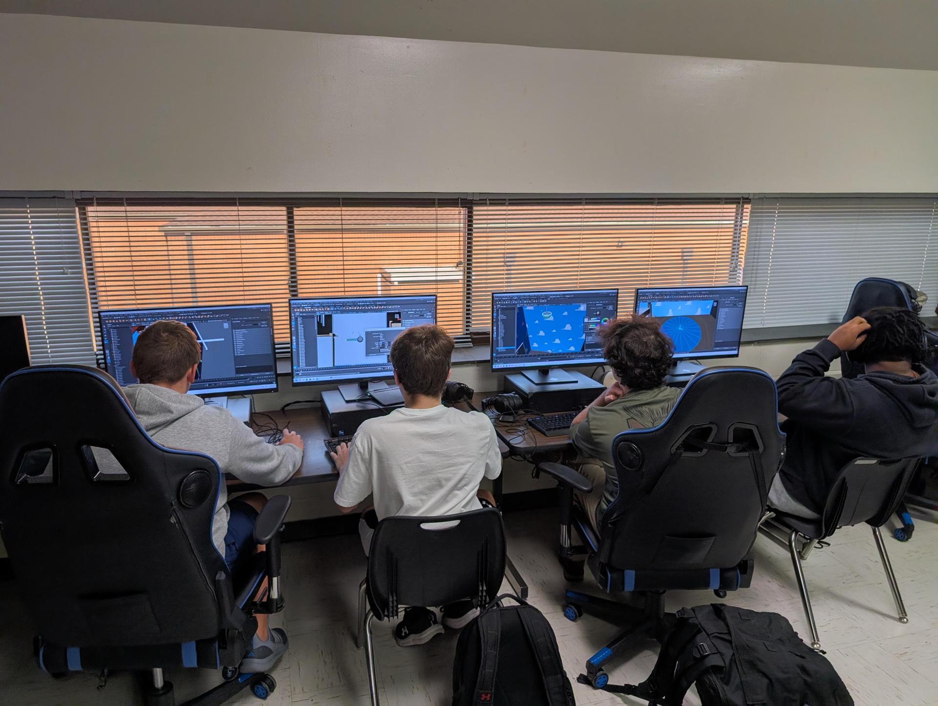 Four students collaborating at computers with multiple monitors displaying designs.
