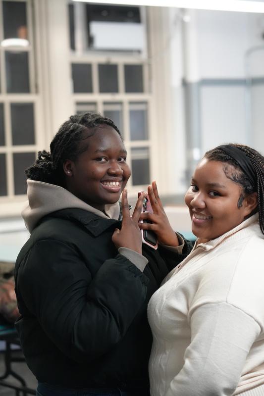 Two smiling young women posing closely together, one with a phone and peace signs, indoors.