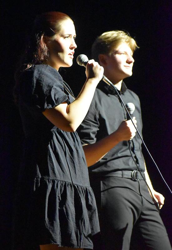 Two performers, one in black, sing on stage with microphones.