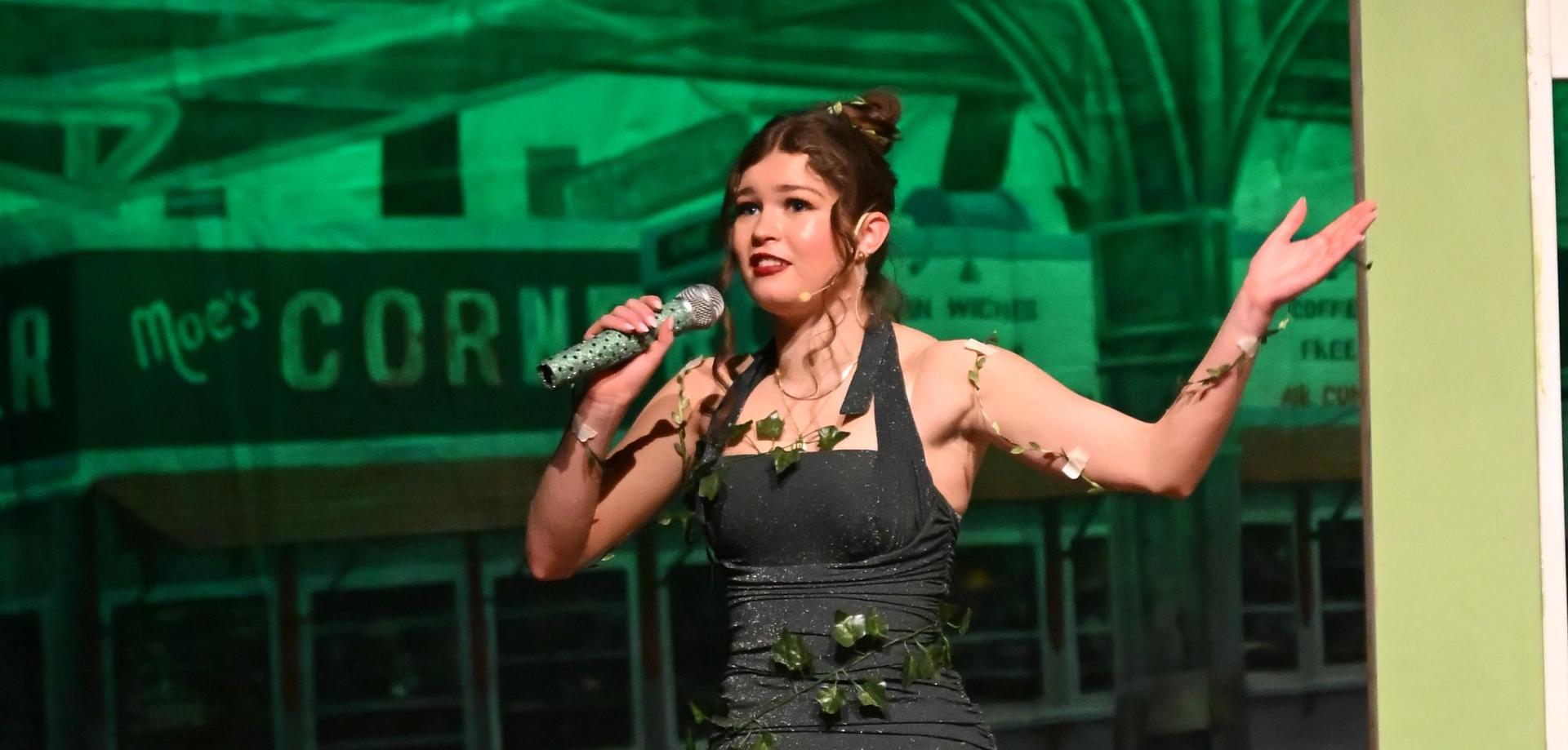 A performer in a black dress adorned with green ivy, singing into a microphone.