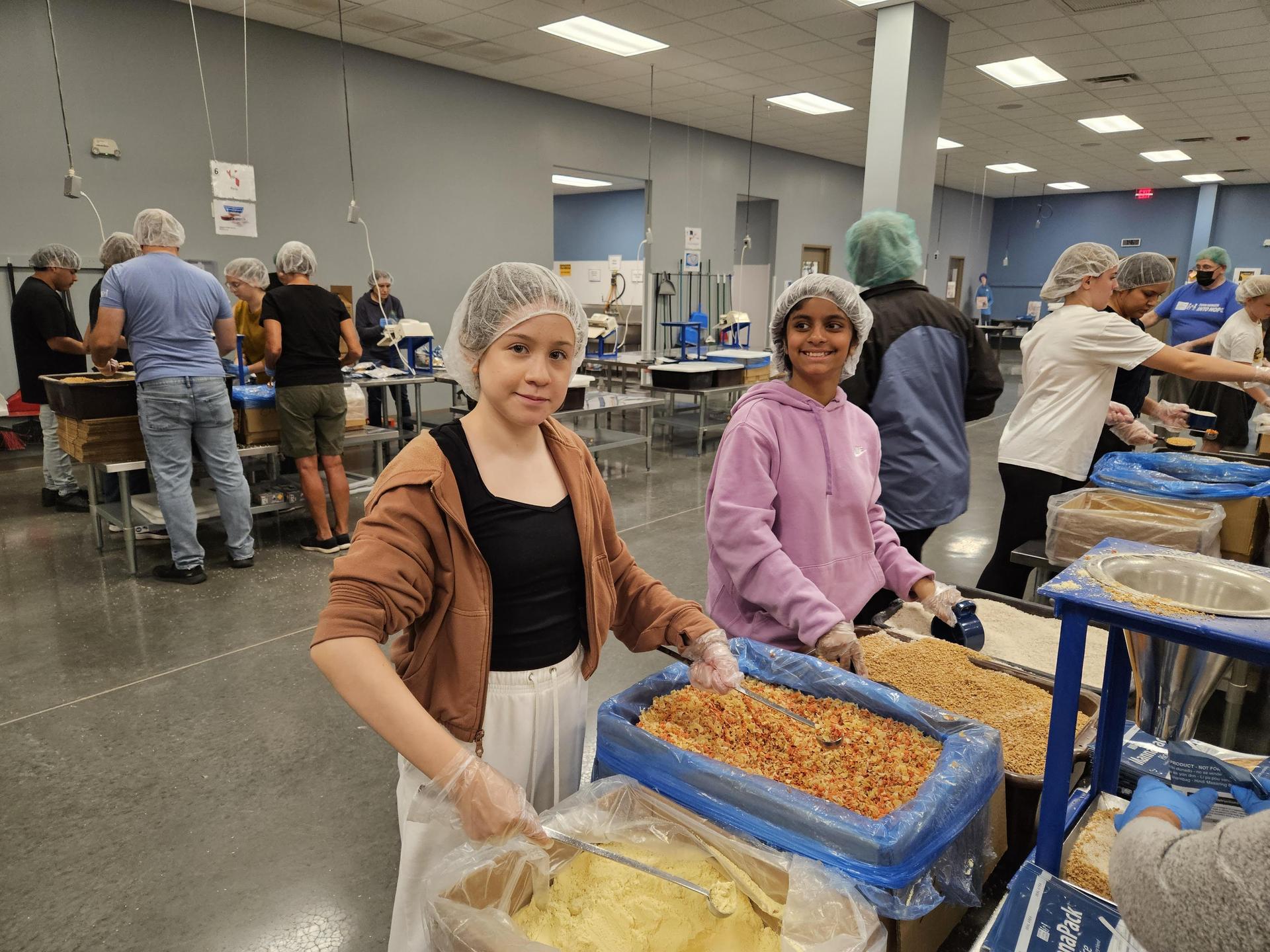 Two young girls volunteering in a food packing event surrounded by people.