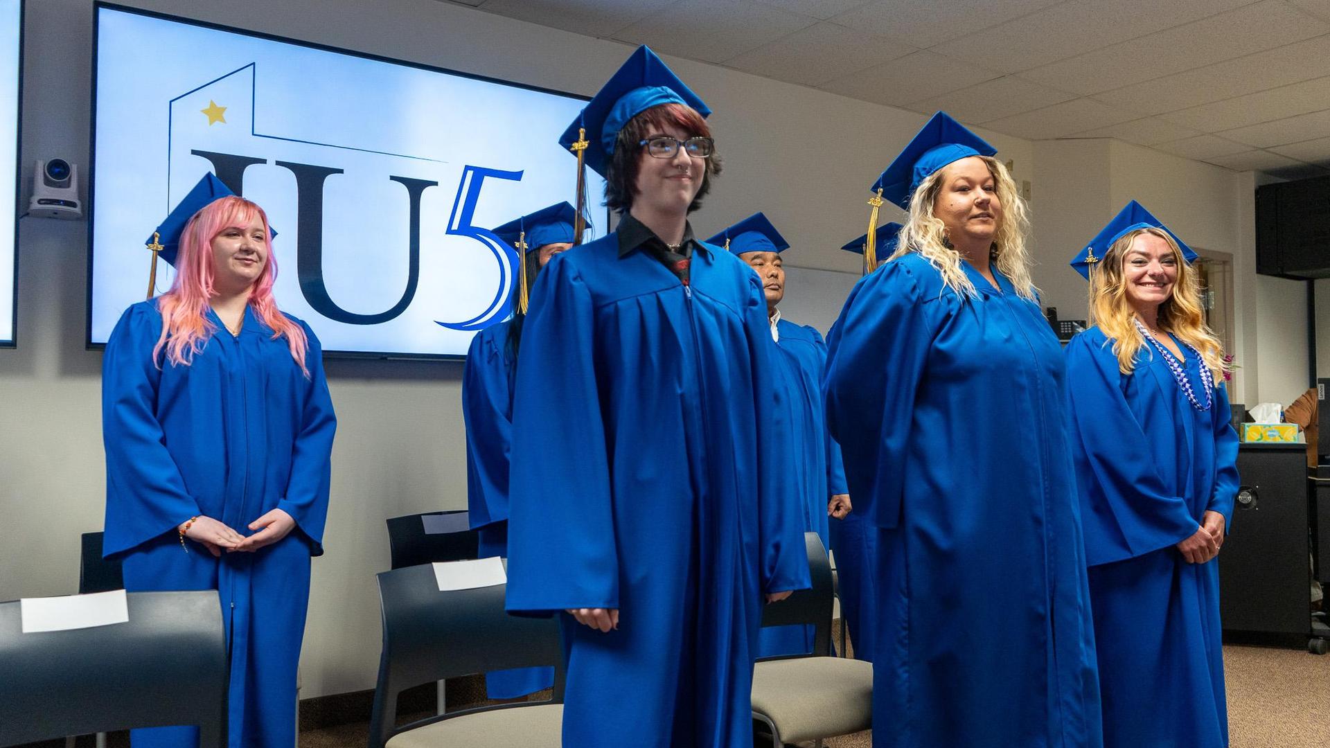 Adult Education graduates in blue caps and gowns standing in a group at their graduation ceremony.