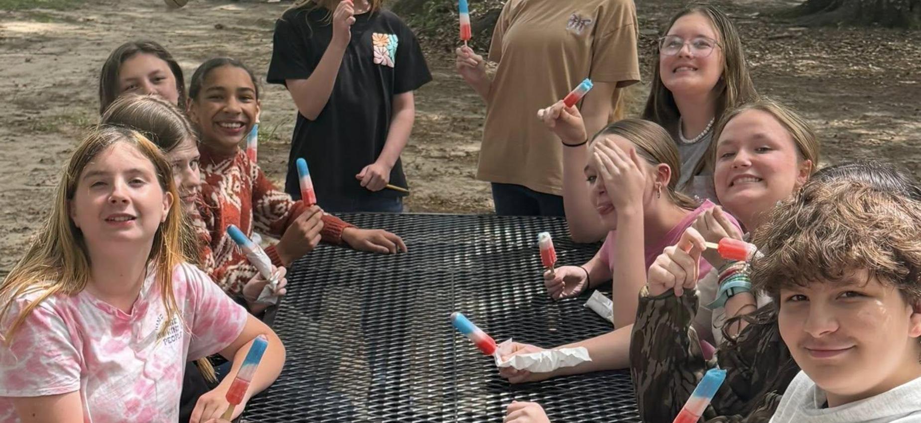 Group of children holding popsicles and sitting at a picnic table outdoors.
