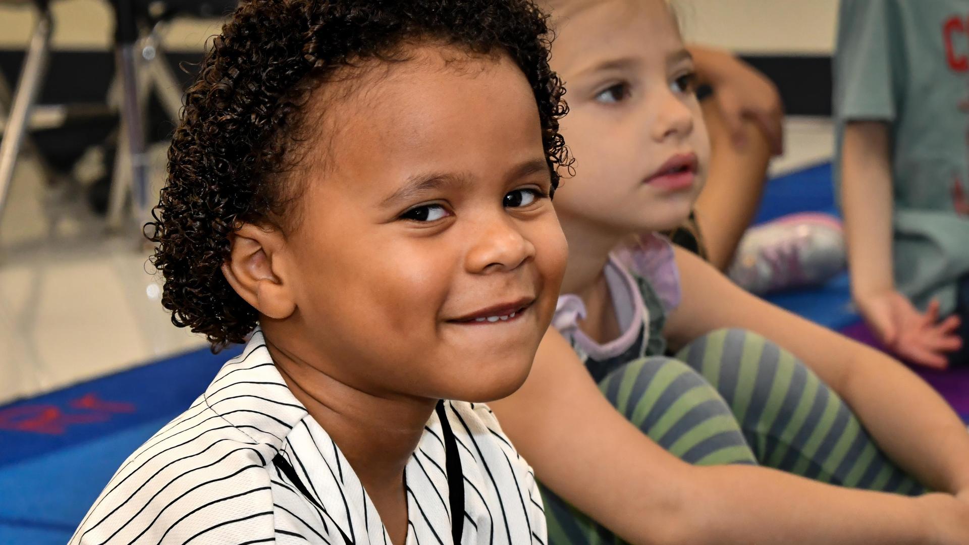 Child with curly hair smiling, sitting among peers in a classroom setting.