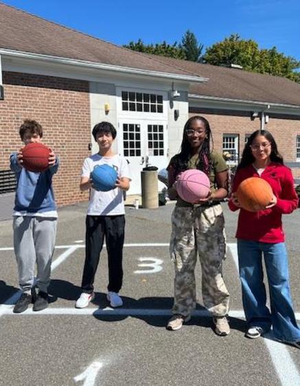 Four students stand outside holding basketballs, ready to play in a schoolyard.