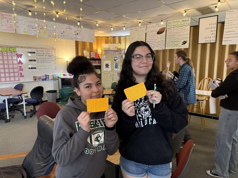 Two girls holding orange cards, smiling in a classroom filled with posters.