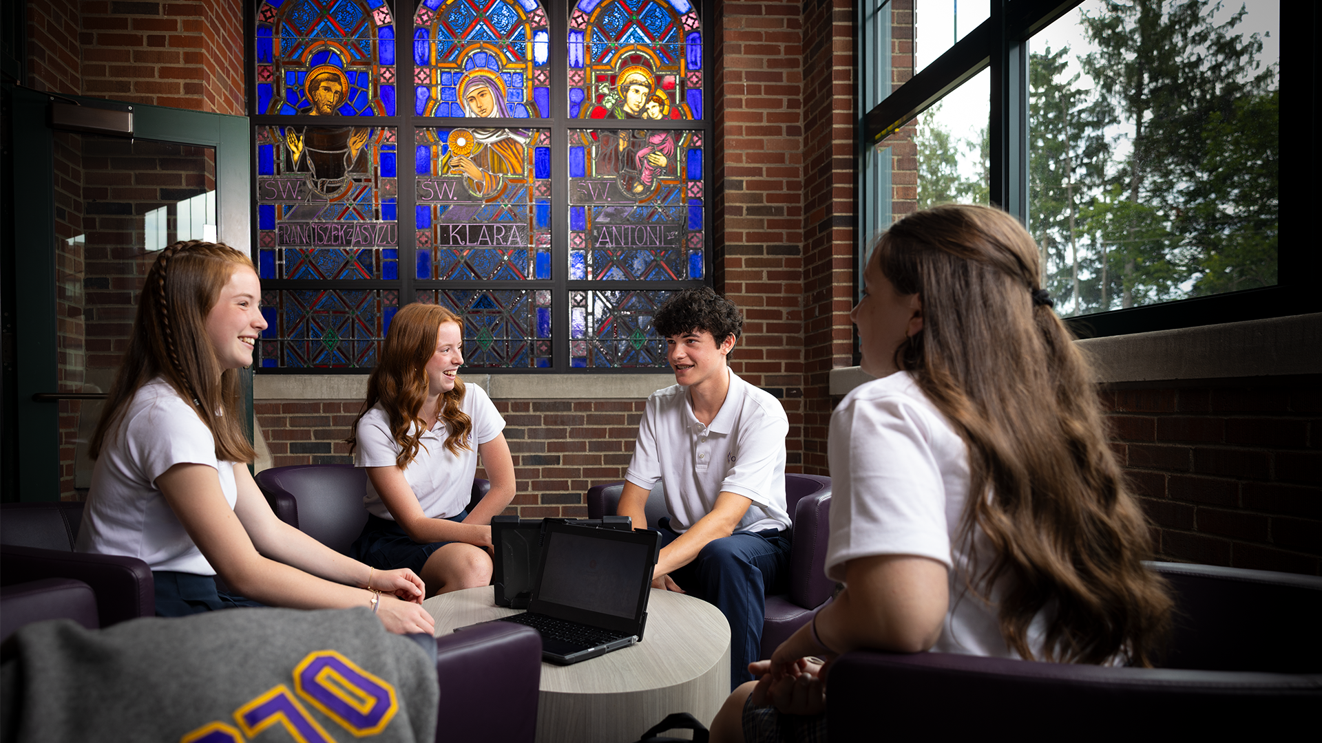 four OLSH students sitting in comfortable chairs talking in front of 3-panel stained glass window