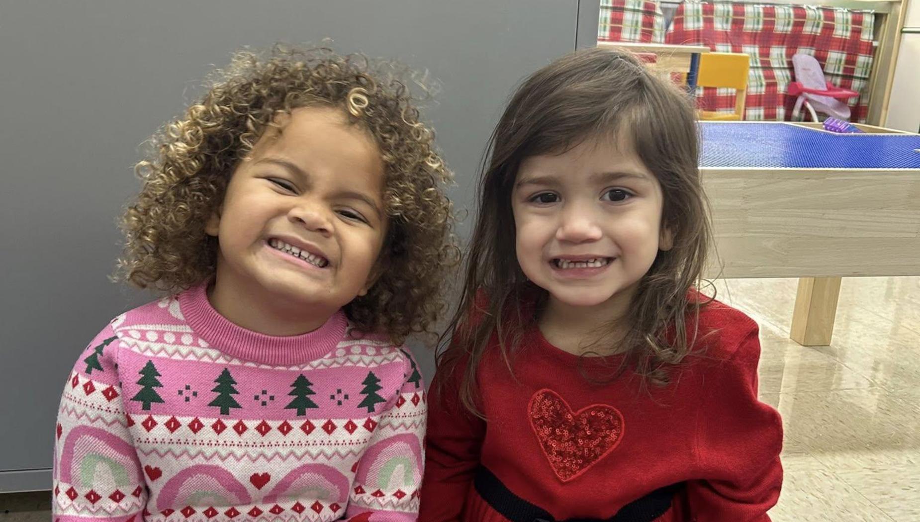 Two girls grinning brightly, one in a festive sweater and the other in a red dress.