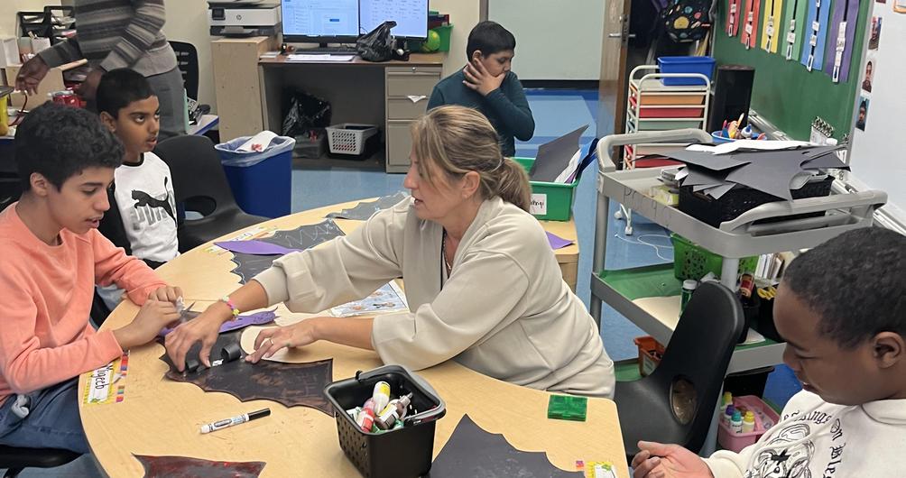 Students and a teacher engaging in craft activities at a table with bat cutouts.