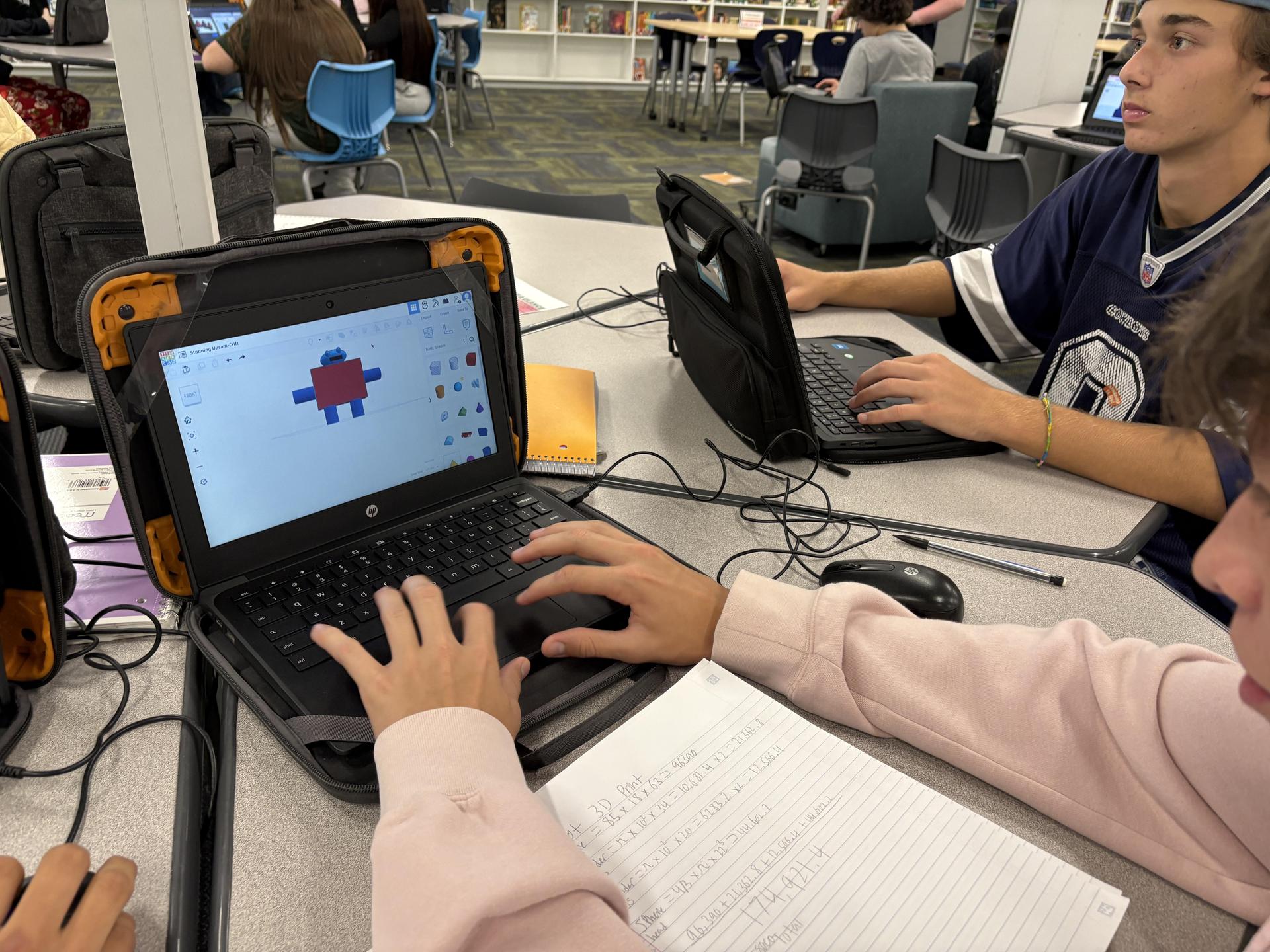 Person using a laptop to create a graphic while seated at a table with a notebook.