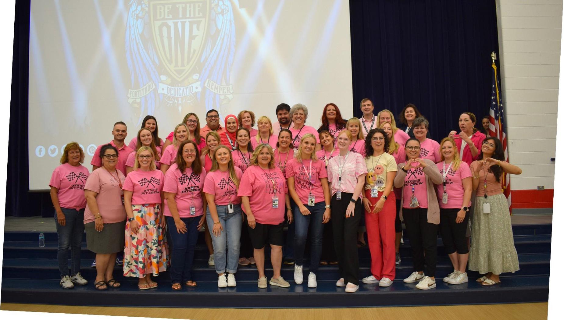 A large group of staff members in pink shirts gathers on stage with a motivational backdrop.