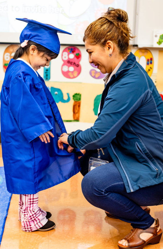 Teacher kneeling to assist student with blue kindergarten graduation robe