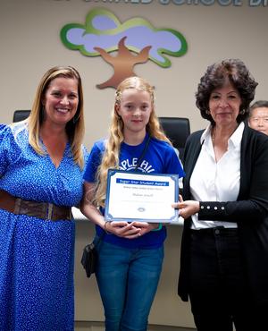 Three people at a school board event, with a girl receiving an award.