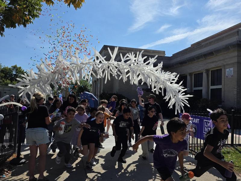 Children and adults celebrating under a colorful confetti archway on a sunny day.