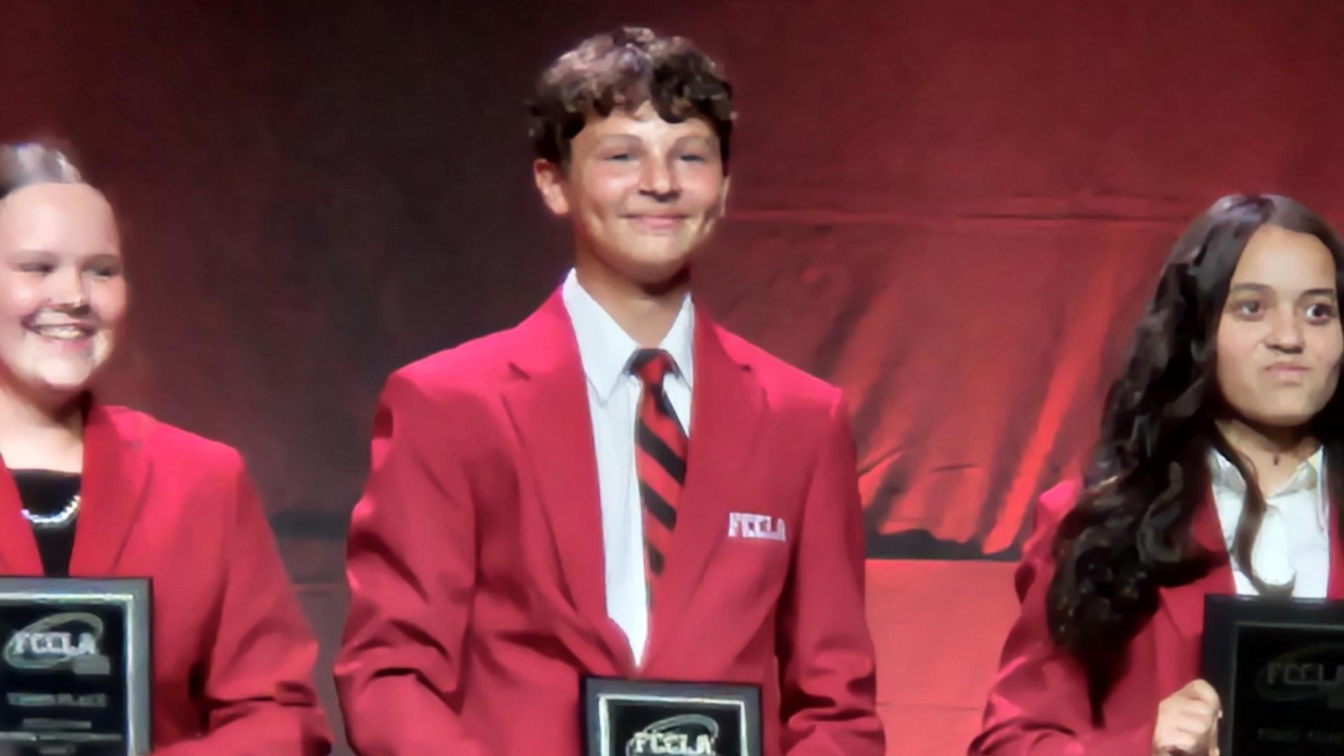 Three individuals in red blazers holding awards on a stage.