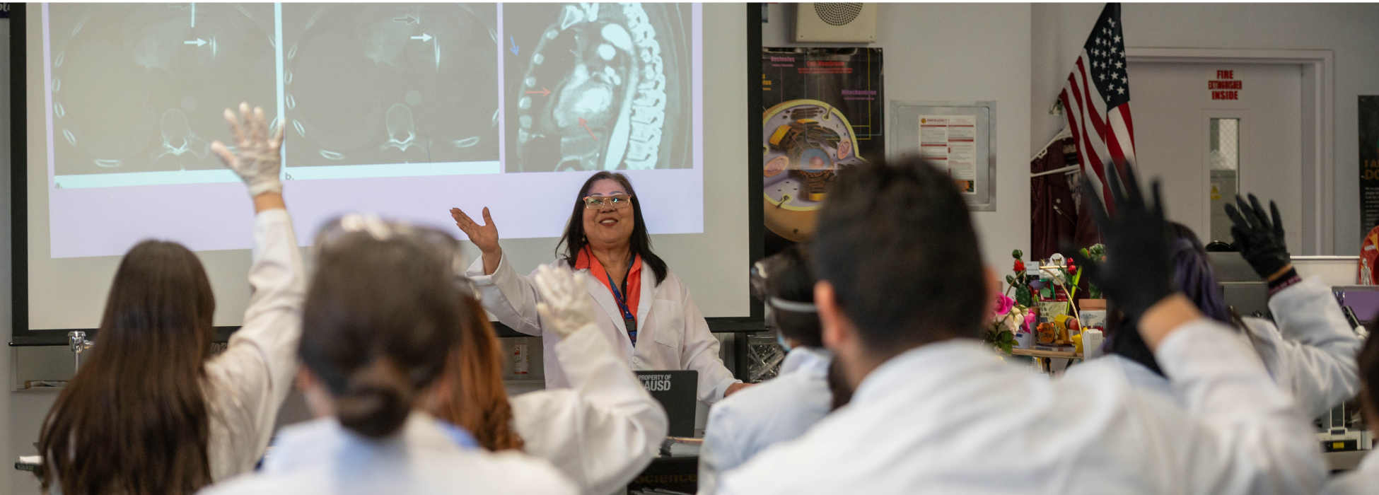 A teacher presents to students in a classroom, engaging with raised hands.