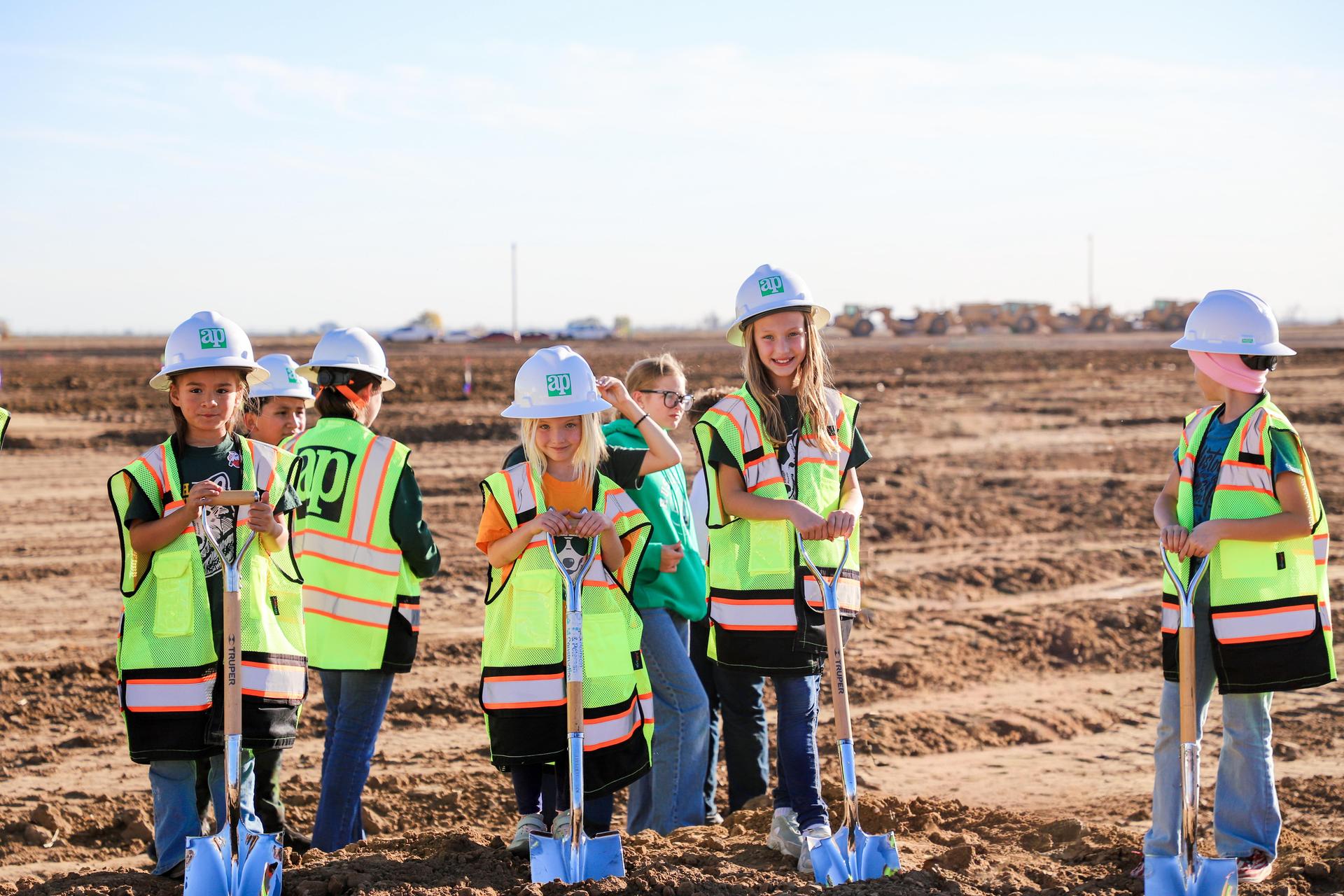 Children dressed in safety gear holding shovels at a construction site.