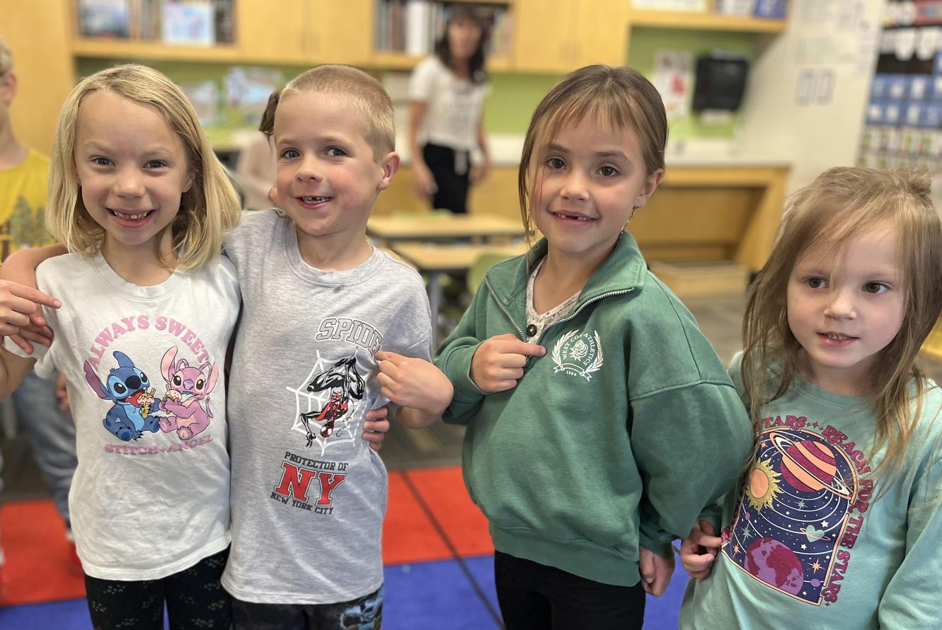 Four children stand on a colorful carpet, pointing at their shirts in a classroom setting.