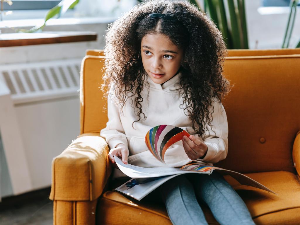 Girl with curly hair sits on an orange couch, reading a magazine.