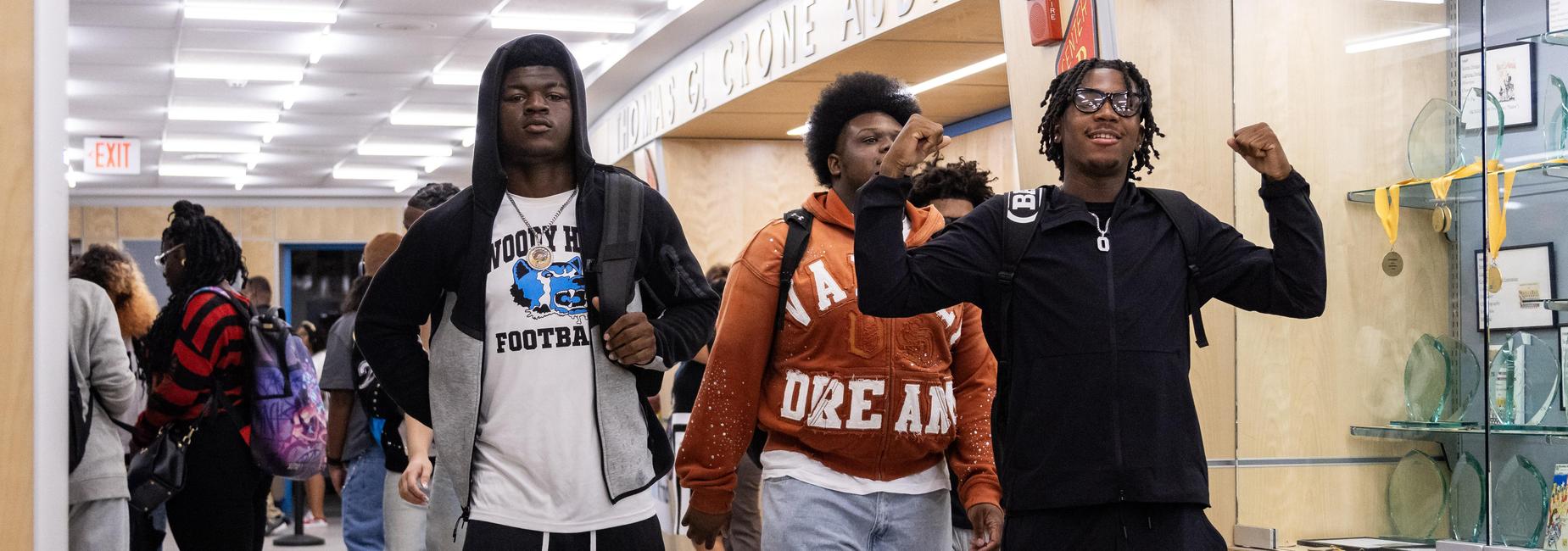 Three high school boys walk through the halls during arrival, with one flexing and smiling