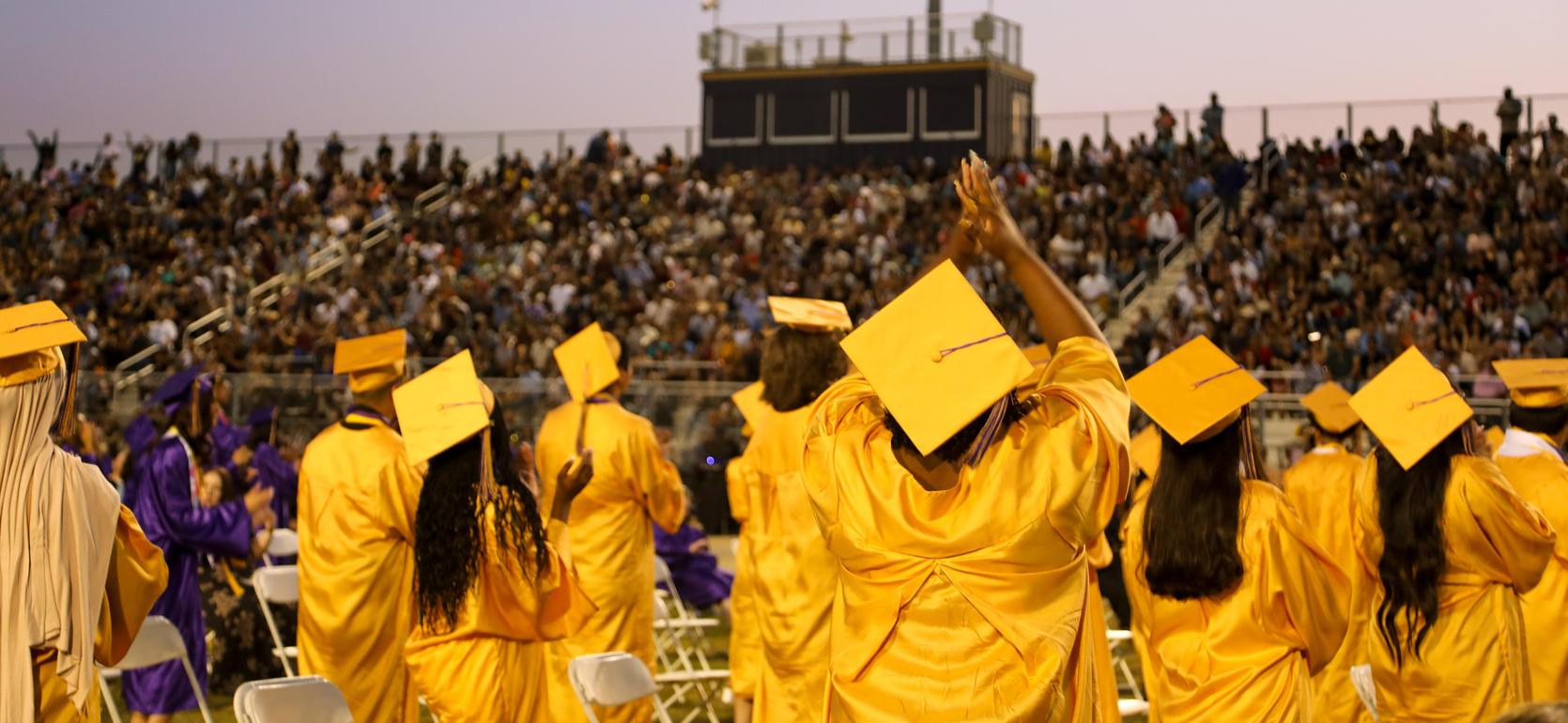 Large crowd of graduates wearing yellow and purple caps celebrating at a graduation ceremony.