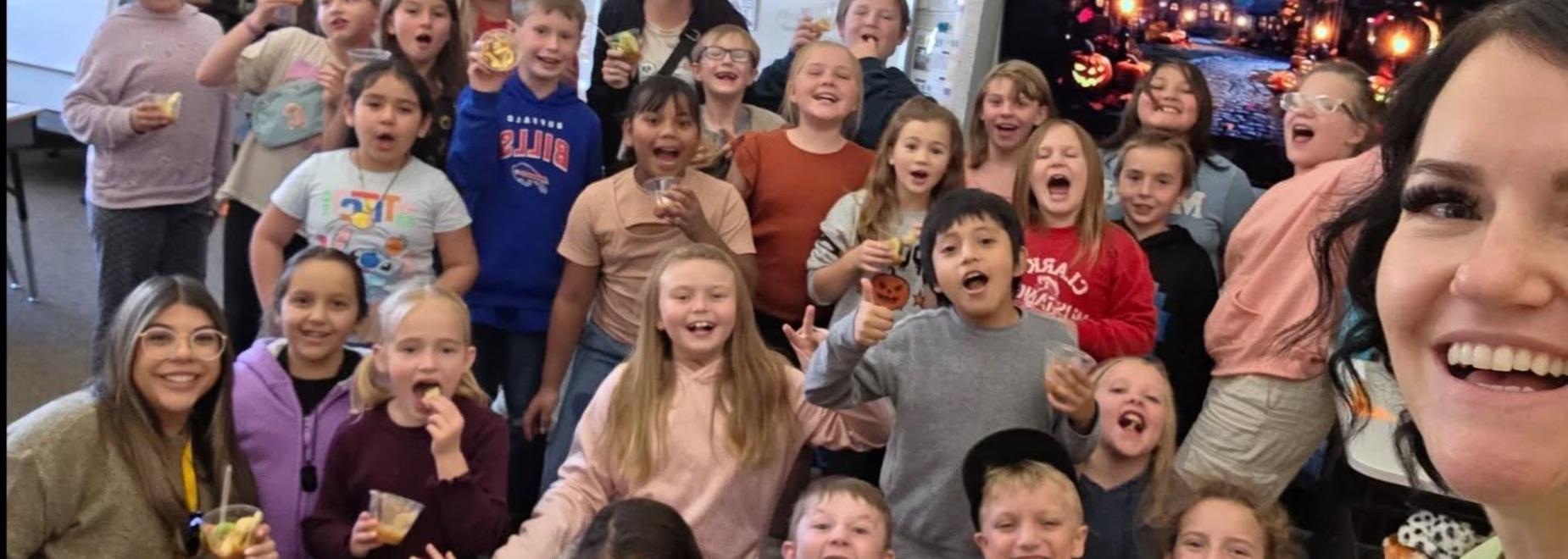 A large group of smiling students posing with snacks and a teacher in a classroom.