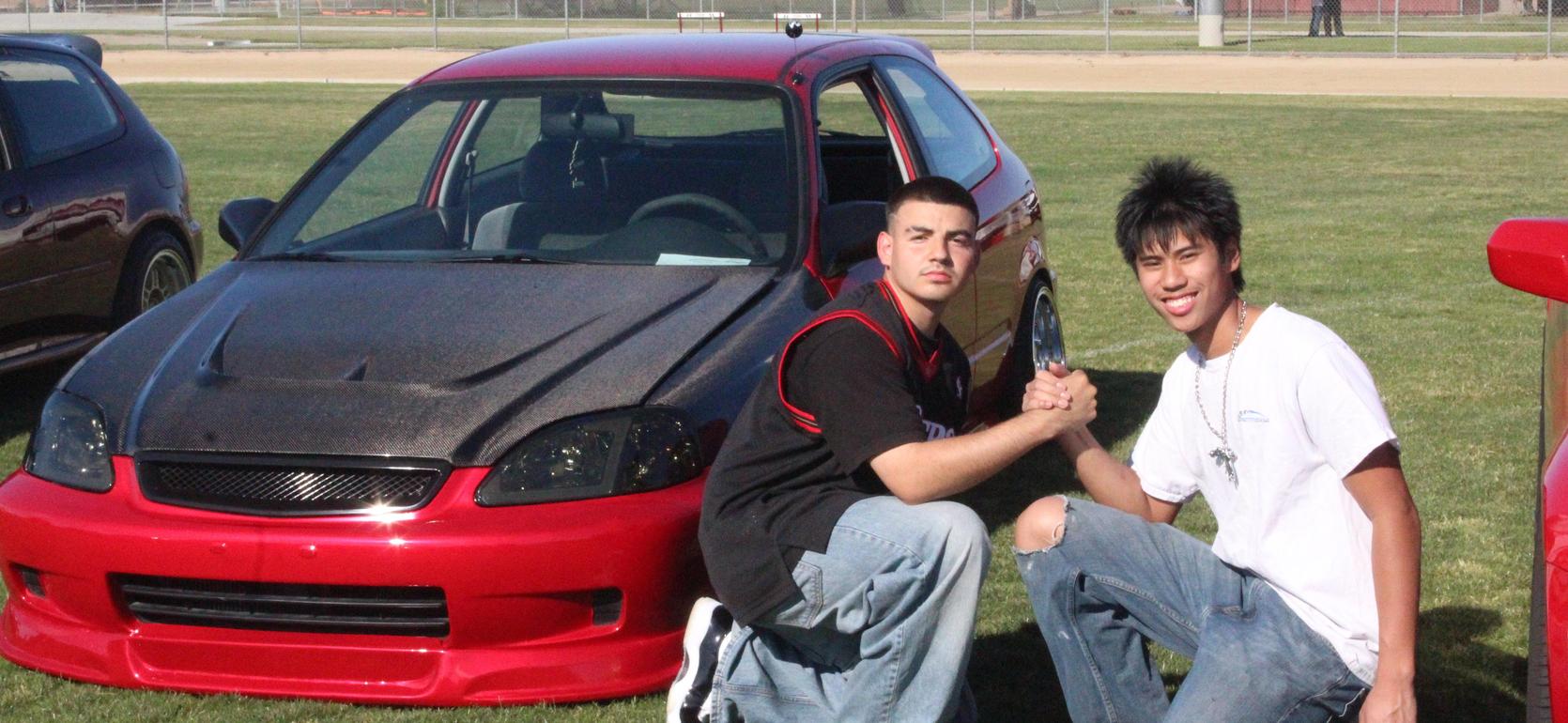 Two young men pose beside a modified red car, holding a trophy in the grass.