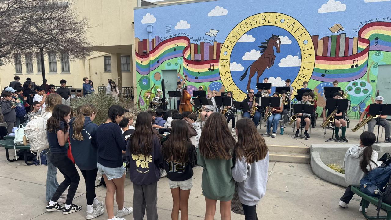 Students gather around a band playing in front of a colorful mural.
