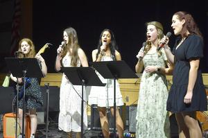 Five young women singing together, one playing an electric guitar.