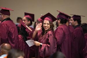 Female graduate in maroon cap and gown beams and waves to the camera while holding her diploma during the recessional portion of the ceremony.