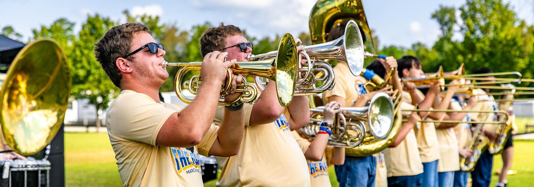 Marching band members playing brass instruments outdoors with sunny background.