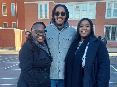 Three smiling people are standing together outdoors in a parking area.