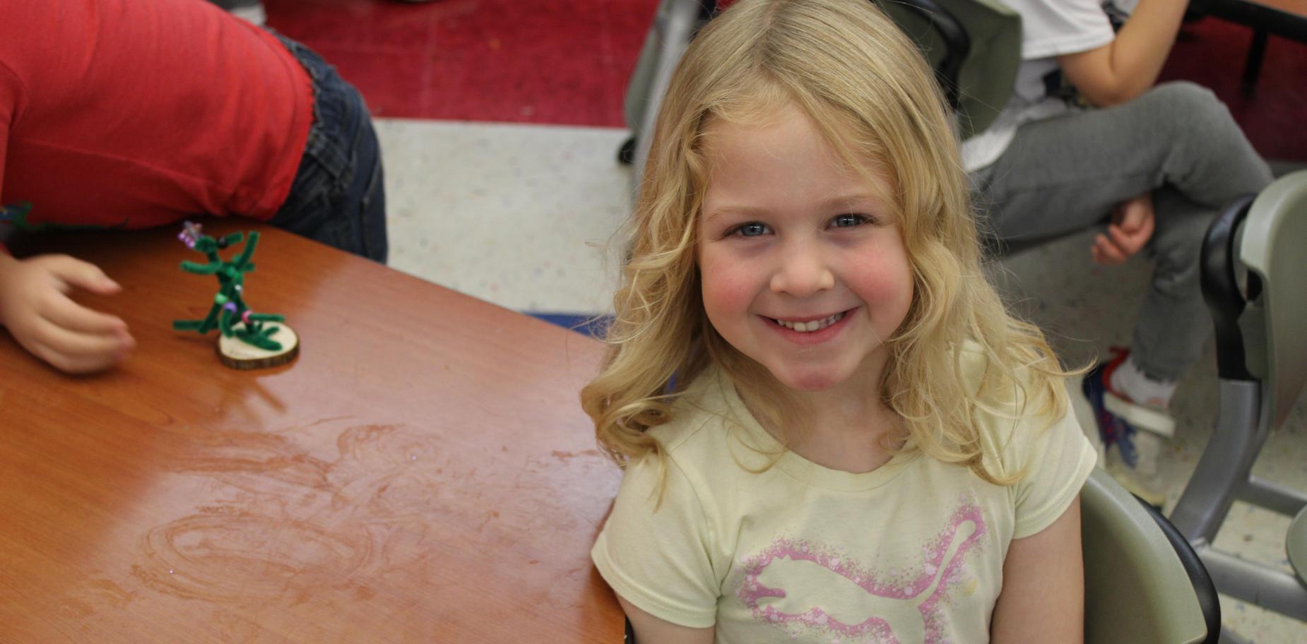 Girl smiling next to a small crafted decoration on a table.
