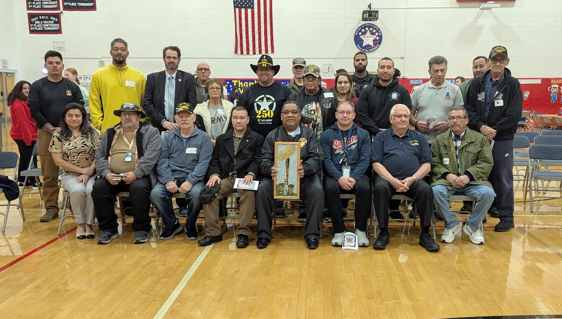 Large group of veterans and attendees posing together in a gym with an American flag.