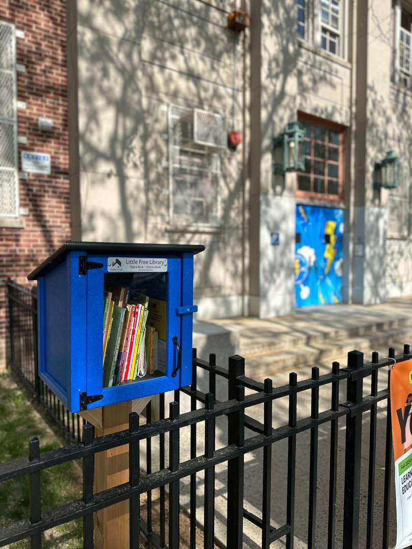 A blue little free library filled with books outside a school building.