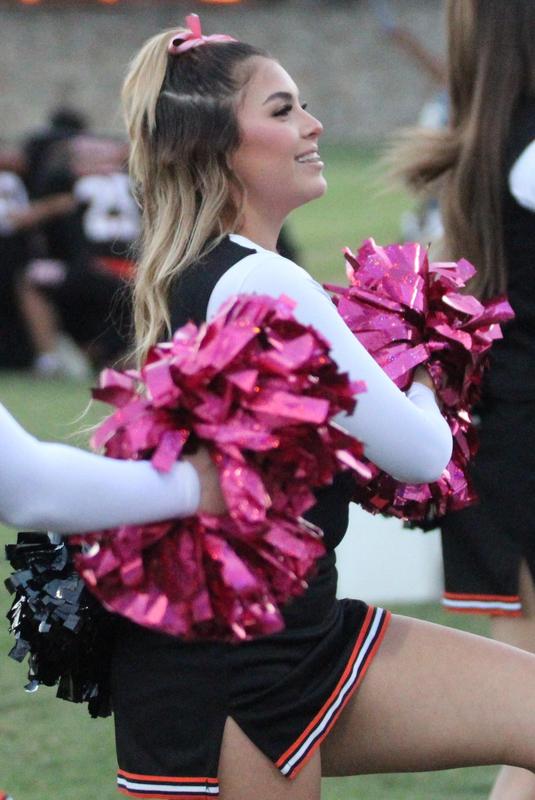 A cheerleader with pink and black pom-poms smiles while performing.