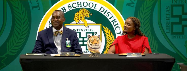 Two individuals seated at a table with a tiger statue, logo in the background.