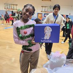 Girl wearing a festive outfit holds a blue box in a gym filled with people.