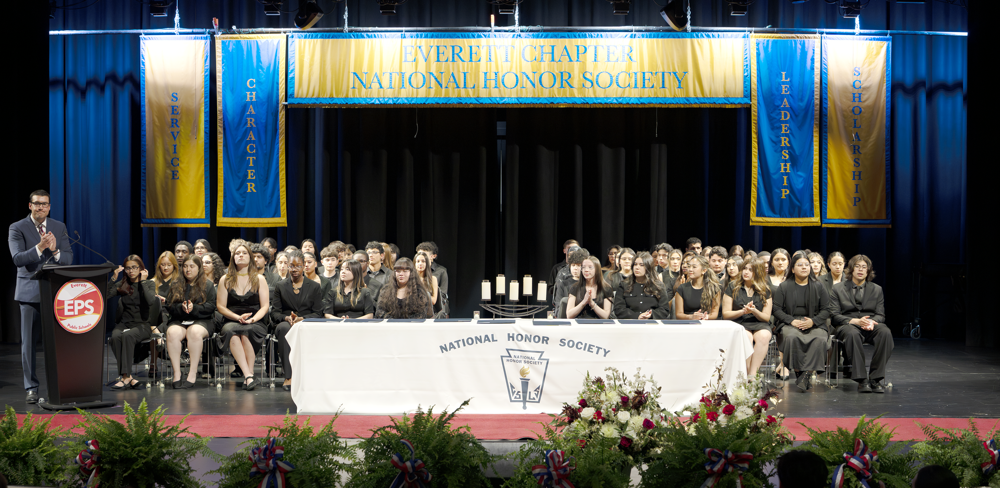 Group ceremony with students in black attire, stage decorations, and a speaker.