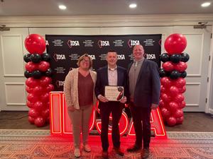 man holding award stands between a female and male with balloon backdrop