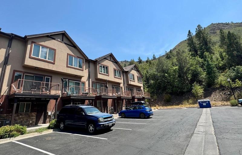 A brown apartment building with cars parked in front and hilly terrain in the background